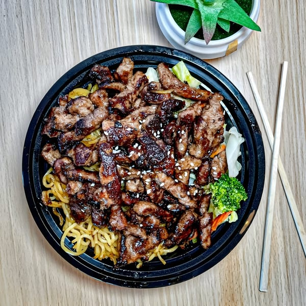 Tokyo Teriyaki's beef teriyaki bowl with noodles, vegetables, and sesame seeds, with chopsticks and a plant.